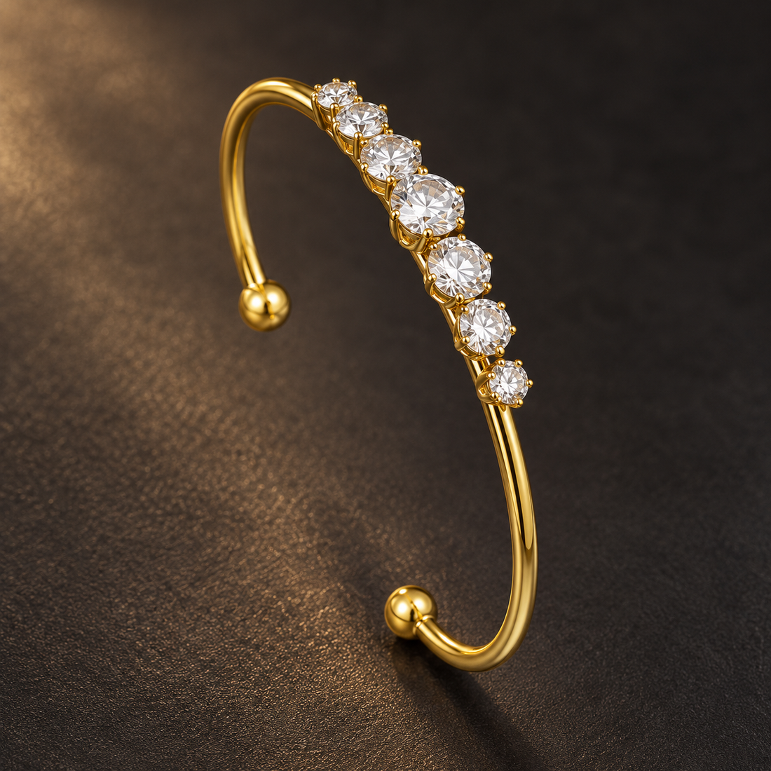 Gold-tone 316L stainless steel cuff bracelet with a row of graduated clear crystals set in prongs and ball-ended open ends, on a dark surface
