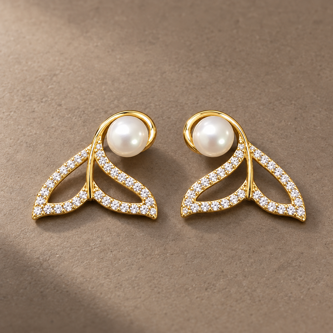 Gold-tone 316L stainless steel earrings in a pair, with a whale-tail shape set with clear stones and a white pearl at the top, on a flat grey surface
