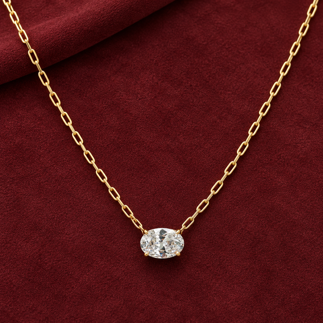 Gold-tone 316L stainless steel necklace with a paperclip chain and an oval clear crystal pendant in a four-prong setting, laid on a dark red cloth