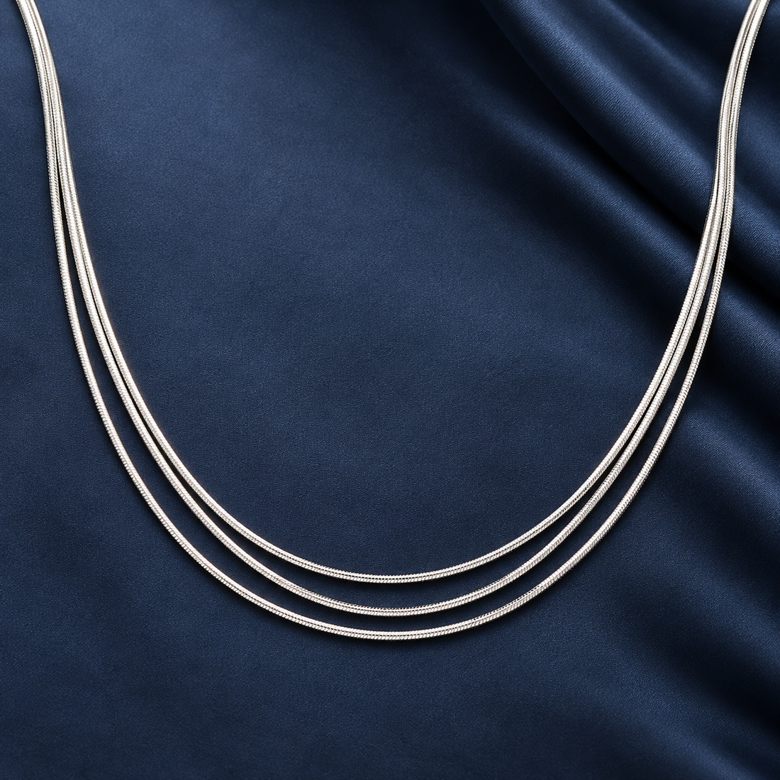 Silver-tone 316L stainless steel three-strand snake chain necklace laid flat on a dark blue fabric, detail view