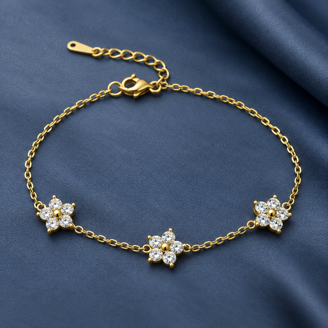 Gold-tone 316L stainless steel chain bracelet with three flower motifs set with clear crystals and a lobster clasp, laid flat on blue fabric