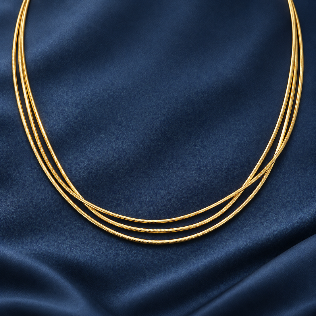 Gold-tone 316L stainless steel necklace of three thin round snake chains laid side by side on a dark blue fabric