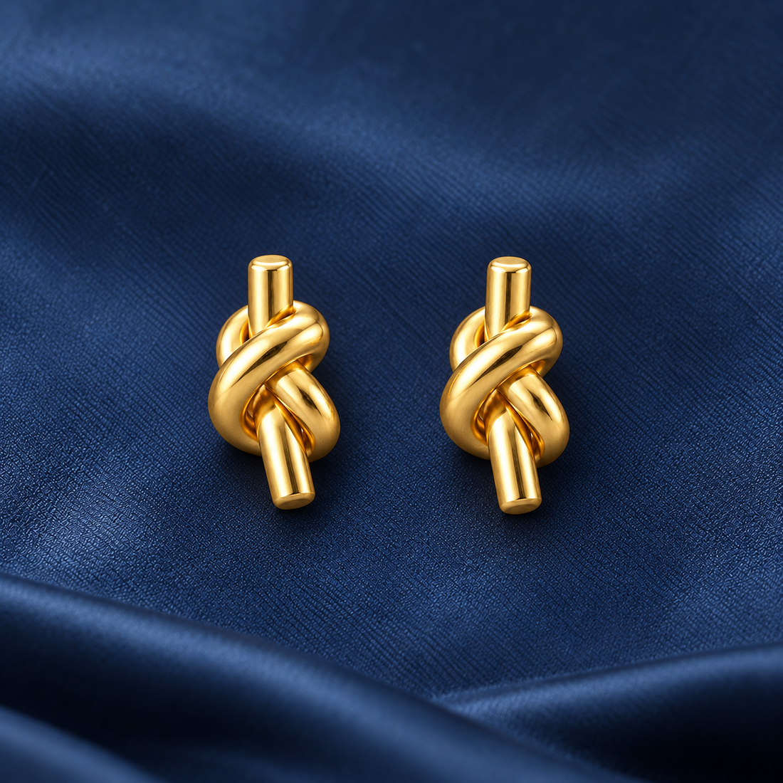 Gold-tone 304 stainless steel pair of earrings with a knotted tube design and straight post backs, shown on a navy fabric