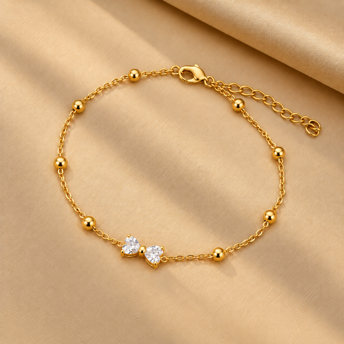 Gold-tone 304 stainless steel bracelet with a fine cable chain, round beads and two heart-cut clear crystals set side by side, laid flat on fabric