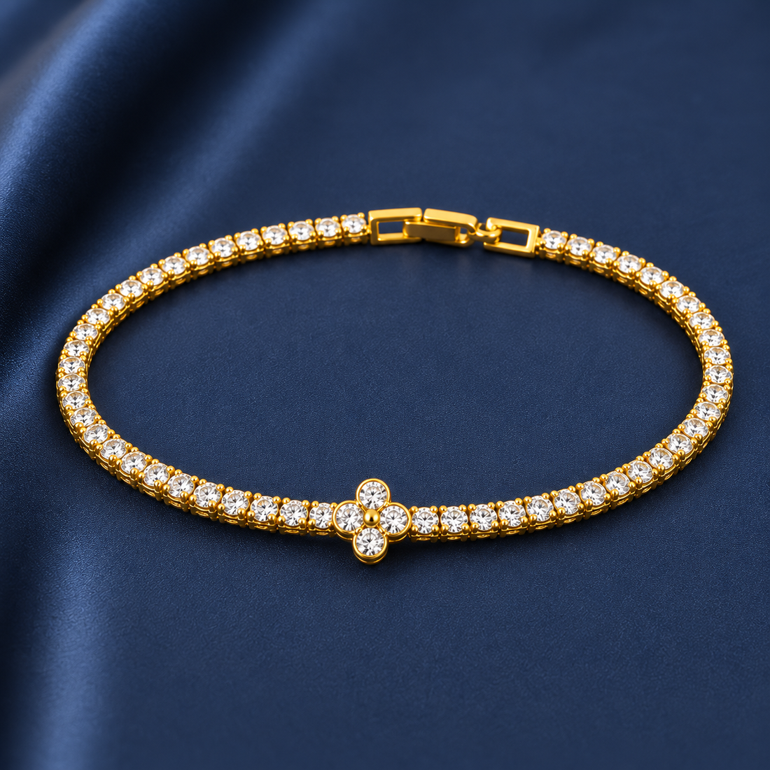 Gold-tone brass tennis bracelet with clear stones in a line and a four-petal flower motif at the centre, laid flat on a navy fabric
