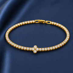 Gold-tone brass tennis bracelet with clear stones in a line and a four-petal flower motif at the centre, laid flat on a navy fabric