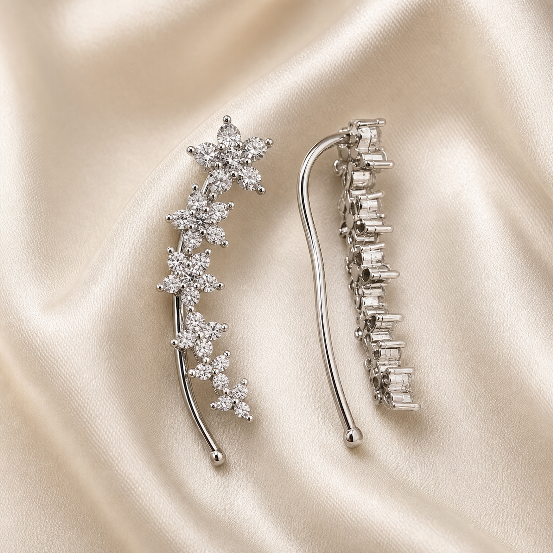 A matched pair of ear-climber earrings, shown from both the decorative face (one earring) and the reverse (the other). The metal is silver-toned with a polished finish. Each earring is a single rigid curved stem of slim wire that hooks over the ear, carrying a graduated line of floral clusters along its outer edge; the reverse shows a series of open box-style prong settings in a stepped row.