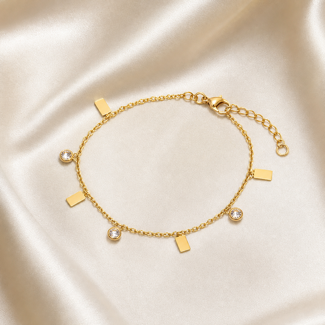 One wrist-worn chain bracelet in yellow-gold-toned metal with a polished finish. It is a single fine cable chain of small oval links carrying dangling charms at irregular intervals; the main chain run is roughly 25 times the width of the largest charm, and a short extender chain of the same link type, about one fifth the main chain's length, terminates in a small ring. A working lobster clasp is present.