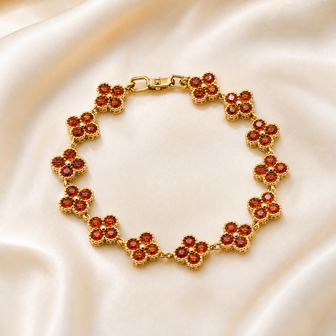 One wrist-worn chain bracelet in yellow-gold-tone metal with a polished finish. It is a single strand of repeating solid-metal quatrefoil (four-lobed clover) stations joined by small round connector links, each station bezel-set with four round faceted red-orange stones arranged one per lobe around a tiny central metal bead; stone colour varies slightly from deep red to orange across stations, with a couple of lobes showing a pale, bright or empty-looking seat.