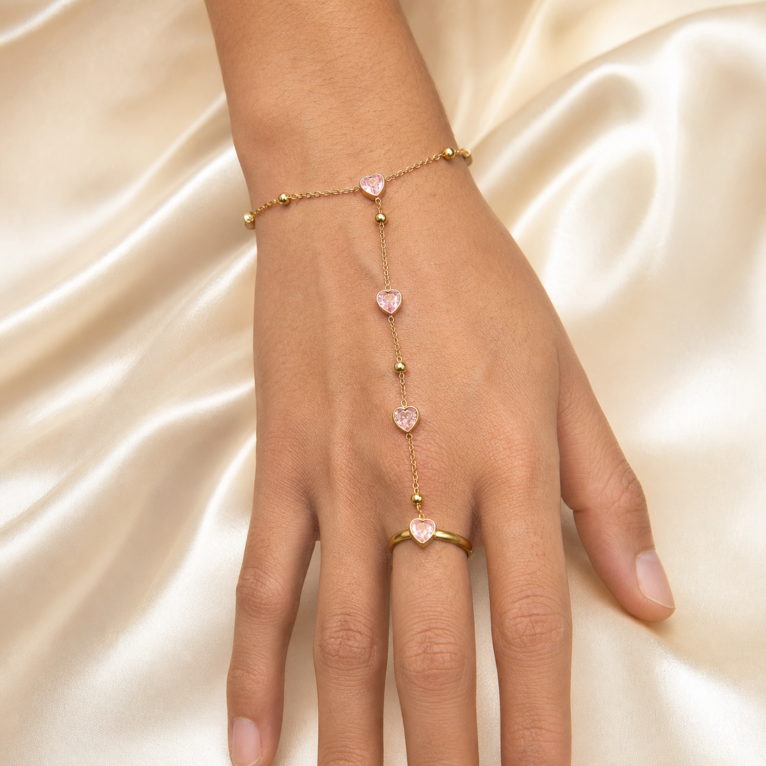 One hand chain (bracelet-to-ring slave bracelet) in yellow-gold-toned metal with a polished finish. The wrist section is a single fine cable chain of fine gauge, carrying two small round polished metal beads and one heart-shaped charm set with a pink heart-cut stone in a bezel-style mounting; the clasp is not visible in the image.