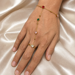 One gold-coloured hand chain (ring-to-wrist bracelet) in polished yellow-gold-tone metal. The wrist section is a single fine gauge cable chain carrying two bezel-set heart-cut stones (one red, one pink) separated along its length by small polished gold spacer beads; its closure is partly obscured and no clasp or extender is resolvable.