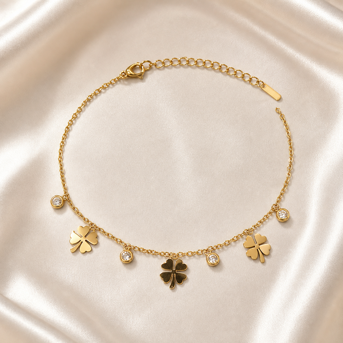 One anklet in yellow-gold-tone metal with a polished finish. It is a single-strand, fine-gauge cable chain with small oval links. The chain run is approximately 30–35 times the width of the largest component (a clover charm); the extender is roughly one-quarter the length of the main chain, terminating in a flat rectangular bar tag. A working lobster clasp is visible. Decoration occupies one central decorated run, with plainer chain toward both the clasp and extender ends.