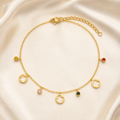 One anklet in yellow-gold-coloured metal with a polished finish, built as a single fine cable chain of small uniform oval links carrying dangling charms along its length. A lobster clasp sits at one end, with a short extender of noticeably larger, chunkier curb-style links, roughly a quarter of the main chain's length, terminating in a plain ring; no separate terminal tag is visible, and the working closure is visible.