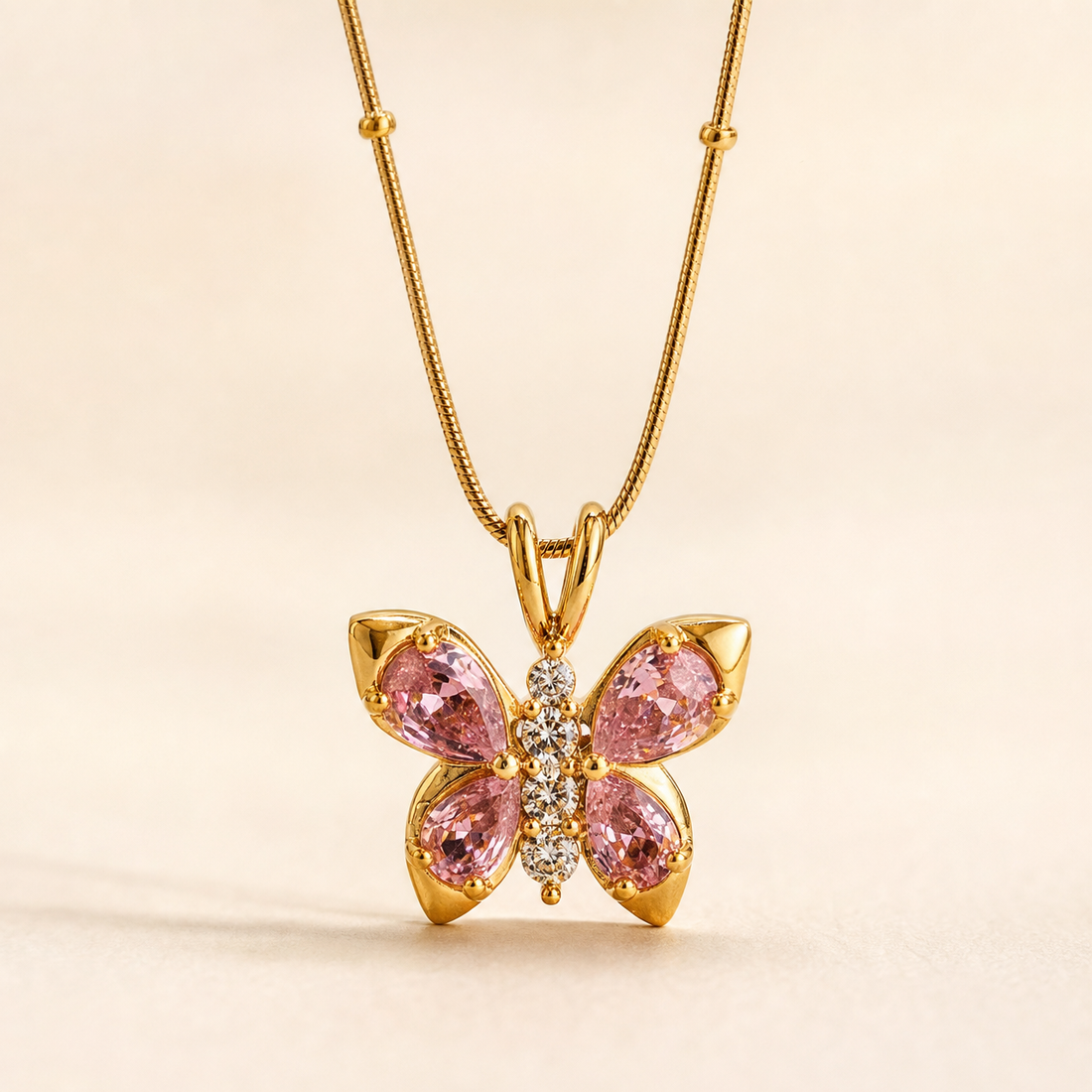 No earrings are present in this image. The visible product is a single pendant necklace: a yellow-gold-tone fine snake chain with small spaced bead stations, suspending a butterfly-shaped pendant through a double-loop bail. The pendant has polished gold-tone metal with four pear-cut pink faceted stones forming the wings in prong settings, and a vertical body of small round clear faceted stones. Pendant drop is roughly one and a half times its widest span.