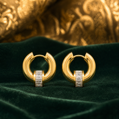 Two ear-worn huggie hoop earrings of matching design, in polished yellow gold with a high-shine finish. Each is a small hinged hoop with a huggie hinge closure; the hoop is chunky and rounded in profile, and the overall drop is roughly one and a half times the hoop's width. Threaded onto each hoop is a single cylindrical barrel element channel-set with a row of clear rectangular baguette-cut faceted stones, free to slide along the hoop rather than fixed.