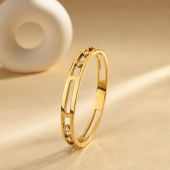One rigid gold-toned bangle with a polished yellow-metal finish. The band is narrow, roughly one-fifteenth of the outer diameter, of even width all round with a slightly rounded flat cross-section. It is pierced with elongated rounded-rectangle cut-outs forming openwork panels: a long central open link flanked by shorter open sections, each holding small round clear colourless stones in raised bezel cups; the stone clusters sit at both ends of the central link, with a couple of stones appearing slightly
