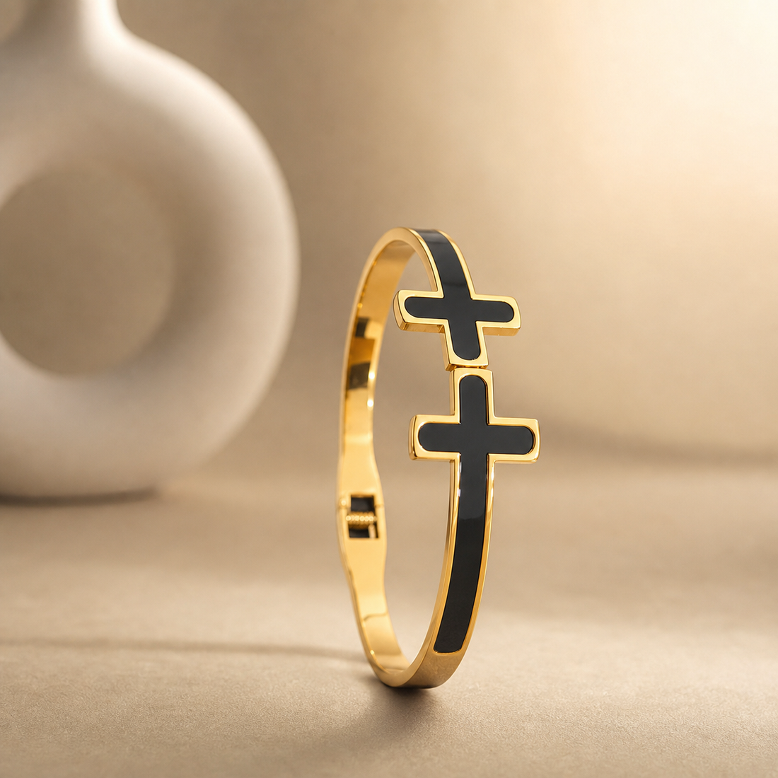 One rigid gold-tone metal bangle with a polished finish. The band is narrow, roughly one-twelfth of the outer diameter in width, of flat cross-section and even width all the way round, each face inlaid flush with black enamel panels running the full circumference. It is a hinged bangle with a box-style clasp opposite the hinge. Two solid-metal cross motifs with rounded arm ends, gold-rimmed and filled with black enamel, sit stacked on the band; the lower cross is larger than the one above it.
