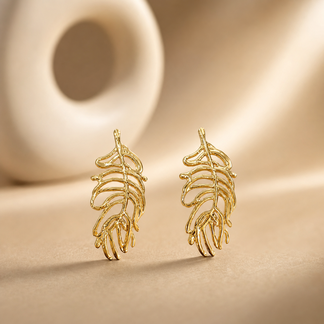 The photograph contains not a kada or bangle but a matched pair of two rigid gold-toned metal ear ornaments, each an openwork feather or leaf motif; no circular band, closure, hinge, gap, terminals or wrist-worn form is present, so band width, cross-section and closure type cannot be reported.