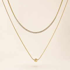 Two separate gold-tone necklaces, polished finish. Necklace one is a single-strand tennis-style chain of small clear round faceted stones, each in an individual gold box/prong mounting linked in a continuous articulated line; stone count is not clearly resolvable at the clasp ends and is withheld, stones are uniform and tightly spaced with no gaps; chain run is roughly 60-70 times one stone width, no extender visible.