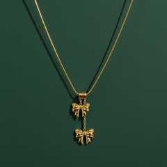 A single yellow-gold-tone necklace with a polished finish. Construction is one fine snake chain of small gauge forming a single strand, carrying a stacked double-bow pendant assembly: a smooth cylindrical barrel bail slides on the chain and supports, by short jump-ring links, a first solid-metal bow charm, from which a short fine chain drop suspends a second bow charm below it.