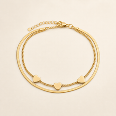 One anklet in yellow-gold-coloured metal with a polished finish. It is a double-strand flexible chain: one strand is a flat, smooth herringbone/snake chain of fine gauge, and the other is a finer rounded curb chain, the two strands joined together at both ends. The main chain run measures roughly twenty-five to thirty times the width of the largest component (the heart charm); a short extender chain of open links, about one eighth of the main chain length, runs from one end.