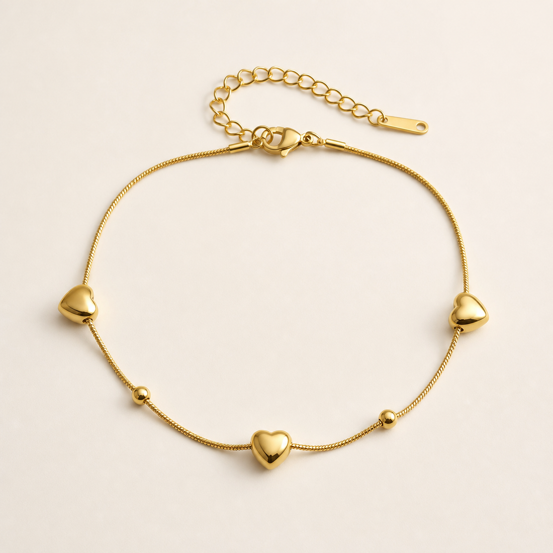 One anklet in yellow-gold-toned metal with a polished finish. Construction is a single strand of fine snake chain with a working lobster-claw clasp; from the clasp extends a curb-link extender chain terminated by one flat perforated bar tag. The main chain run is approximately thirty-five times the width of the largest component, a puffed heart, and the extender is roughly one quarter of the main chain length. Decoration is spaced along the strand rather than continuous, with plain chain between stations.