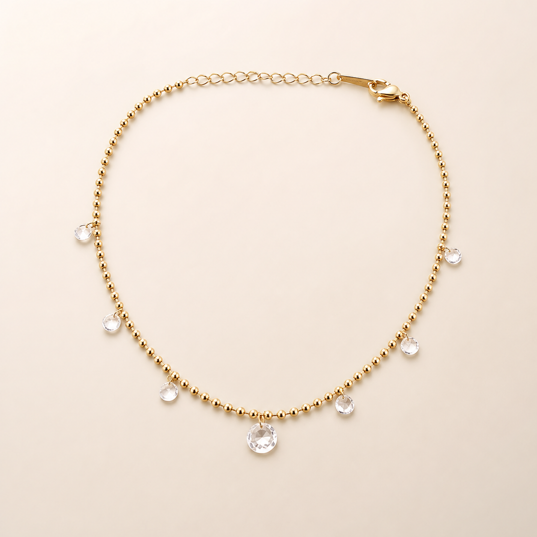 One anklet only: a single flexible yellow-gold-tone polished chain in a fine small-ball/bead construction, with one short oval-cable-link extender section and one visible working lobster-claw closure; no terminal tag is clearly resolved. The main chain run is approximately 30 times the width of the largest dangling component, and the extender is roughly one-eighth of the main chain length.