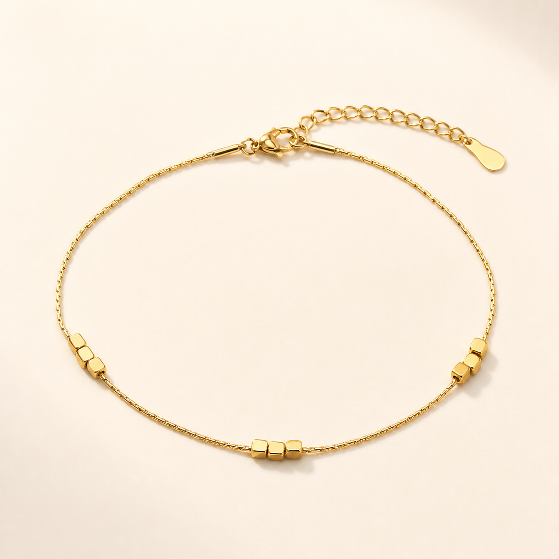 One anklet. Yellow-gold-toned metal with a polished finish, constructed as a single fine snake-type chain of very fine gauge, unbranched, with no second strand. The chain run is approximately 70 times the width of its largest components (the cube beads). A lobster-claw clasp provides a working closure, joined to a curb-link extender chain roughly one sixth the length of the main chain, terminated by a single small flat oval drop tag.