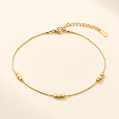 One anklet. Yellow-gold-toned metal with a polished finish, constructed as a single fine snake-type chain of very fine gauge, unbranched, with no second strand. The chain run is approximately 70 times the width of its largest components (the cube beads). A lobster-claw clasp provides a working closure, joined to a curb-link extender chain roughly one sixth the length of the main chain, terminated by a single small flat oval drop tag.