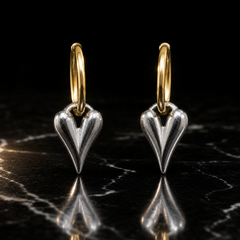 Two earrings of one design, with polished two-tone metal: a small gold-coloured huggie-hinge hoop (fine round wire, about 1 mm gauge, roughly 12 mm across) carrying a silver-coloured solid-metal elongated heart. Drop is approximately twice the widest part, the heart. Two sections articulate: the hinged hoop and the heart, swinging where the hoop passes through the heart's integrated top loop.
