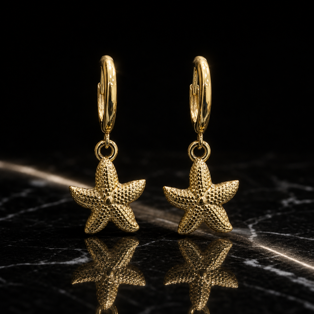 Two earrings, one design: yellow-gold-toned metal with polished fittings and a granular hammered texture on the motifs. Each earring has a small huggie-hinge hoop of medium gauge, with overall drop approximately twice the widest starfish-arm span. From each hoop a single jump ring mounts one solid five-arm starfish dangle, textured across its surface with a small raised central dot, swinging freely at the jump ring. No stones, pearls, discs or tassels are present.