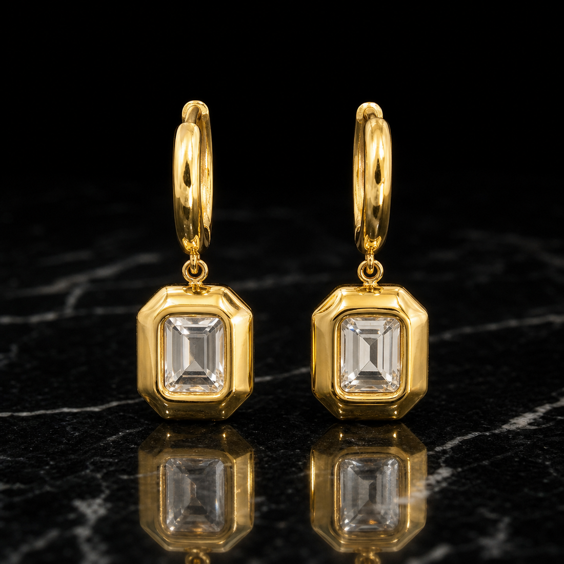 Two earrings of one design in yellow-gold-tone metal with a polished finish. Each has a huggie hinge hoop fitting of short length and medium gauge, with an overall drop about twice the pendant's widest part. From the hoop, a small jump ring articulates a single rigid section: an octagonal rectangular frame pendant. Set within each frame is one clear rectangular step-cut faceted element in a bezel mounting, centred in a solid polished border.