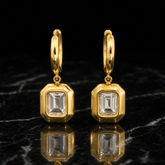 Two earrings of one design in yellow-gold-tone metal with a polished finish. Each has a huggie hinge hoop fitting of short length and medium gauge, with an overall drop about twice the pendant's widest part. From the hoop, a small jump ring articulates a single rigid section: an octagonal rectangular frame pendant. Set within each frame is one clear rectangular step-cut faceted element in a bezel mounting, centred in a solid polished border.