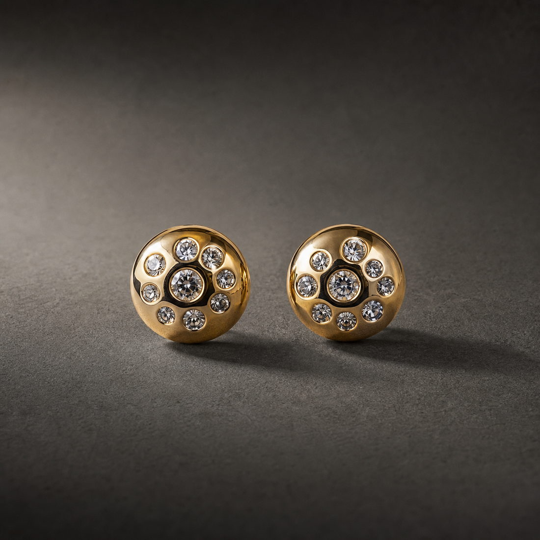 Two ear-worn stud earrings of one design. Each is a polished yellow-gold domed disc, round in outline, its surface flush-set with clear round faceted stones: one larger central stone surrounded by smaller graduated stones scattered across the dome, all in smooth bezel-style recessed settings; exact stone count is not fully resolvable. The earrings are rigid, single-section studs with no articulation or dangling parts; overall depth is roughly equal to the disc width.