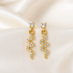 A matched pair of drop earrings, two items total, in yellow-gold-toned metal with a polished finish. Each earring comprises a stud top holding one round clear faceted element in a four-prong setting, with a small jump ring beneath suspending a free-swinging pendant; the pendant is a single wavy, zigzag or lightning-bolt-shaped bar of gold-toned metal whose front face carries a continuous field of small clear round-set stones (individual count not reliably separable) ending in a pointed lower tip.