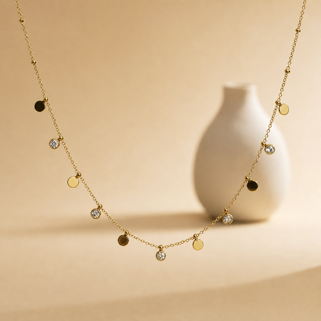 One gold-tone station necklace of a single fine cable chain with polished finish, carrying alternating dangling charms and small round bead stations spaced along the chain. The chain run is roughly 30 times the width of the largest disc component; no extender is visible, and the chain ends leave the frame, so the closure is not visible and clasp and extender type cannot be confirmed.