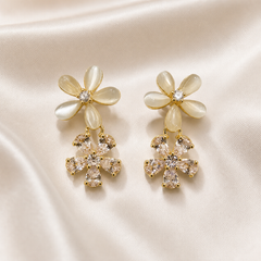 A matched pair of drop earrings, quantity two, in gold-tone polished metal. Each earring comprises two flower motifs joined vertically: an upper flower and a slightly smaller lower flower suspended beneath it. The upper flower has five elongated oval petals of creamy opalescent material with a soft translucent sheen, mounted in gold-tone bezels radiating from a centre holding one small round clear faceted stone in a prong setting.