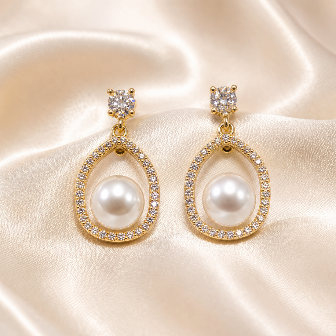 A matched pair of drop earrings, two pieces total. Yellow-gold-tone metal with a polished, bright finish. Each earring consists of a stud top holding one round clear faceted stone in a four-prong basket setting, connected by a short articulated link of two small metal rings to a rigid open teardrop-shaped frame. The frame's outer edge is continuously pavé-set with small clear faceted stones along its full length.