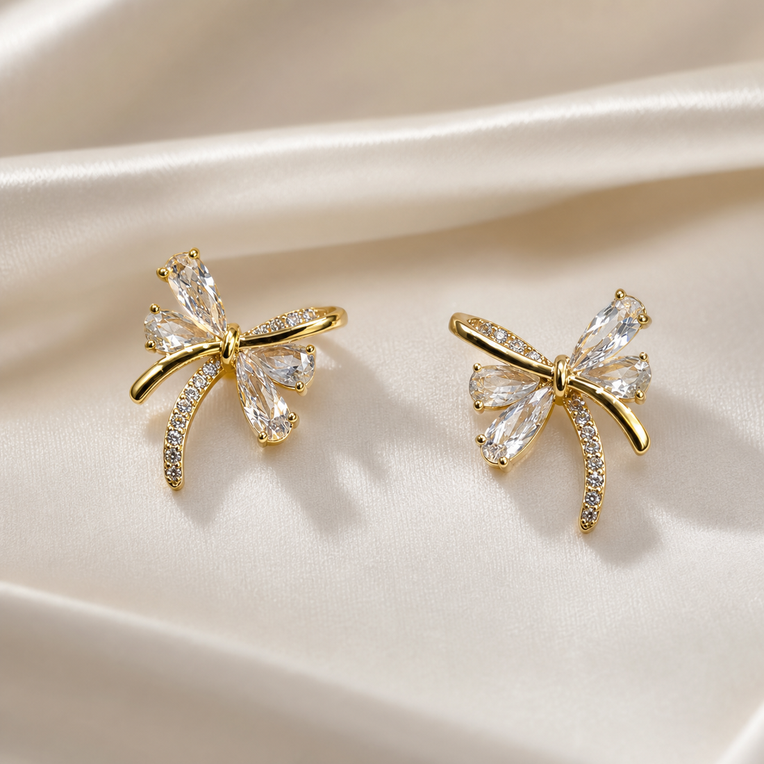 A matched pair of two bow-shaped stud earrings in gold-tone metal with a polished, bright finish. Each earring is a single cast bow motif with a small knotted centre from which four loops and two trailing ribbon ends radiate; there is no chain, strand, clasp or extender, the reverse fittings being partly obscured.