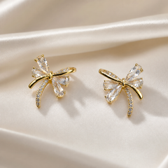 A matched pair of two bow-shaped stud earrings in gold-tone metal with a polished, bright finish. Each earring is a single cast bow motif with a small knotted centre from which four loops and two trailing ribbon ends radiate; there is no chain, strand, clasp or extender, the reverse fittings being partly obscured.