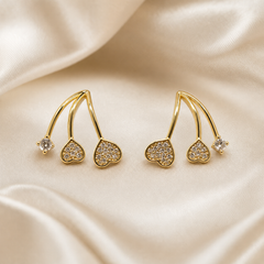 A matched pair of two ear-climber style earrings in polished yellow-gold-toned metal with a smooth reflective finish. Each earring comprises three thin curved solid-metal stems joined at a shared base, each stem terminating in a distinct decorative element: a small rounded heart motif, a larger rounded heart motif, and a single round clear faceted stone.