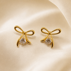 A matched pair of two stud earrings in polished yellow gold-tone metal with a high-shine reflective finish. Each earring is a single solid cast metal motif in the form of a tied ribbon bow, formed from smooth tubular wire: two rounded loops crossing at a central knot with two tapered ribbon tails descending beneath it.