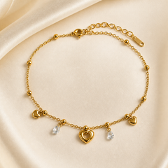 One anklet in gold-coloured metal with a polished finish. It is a single flexible strand of fine cable chain interspersed with small polished round bead stations, with one slightly thicker extension run of larger oval links at the clasp end. The chain run is roughly 25 times the width of the largest component, the central open-heart charm, and the extender is about one sixth of the main chain.