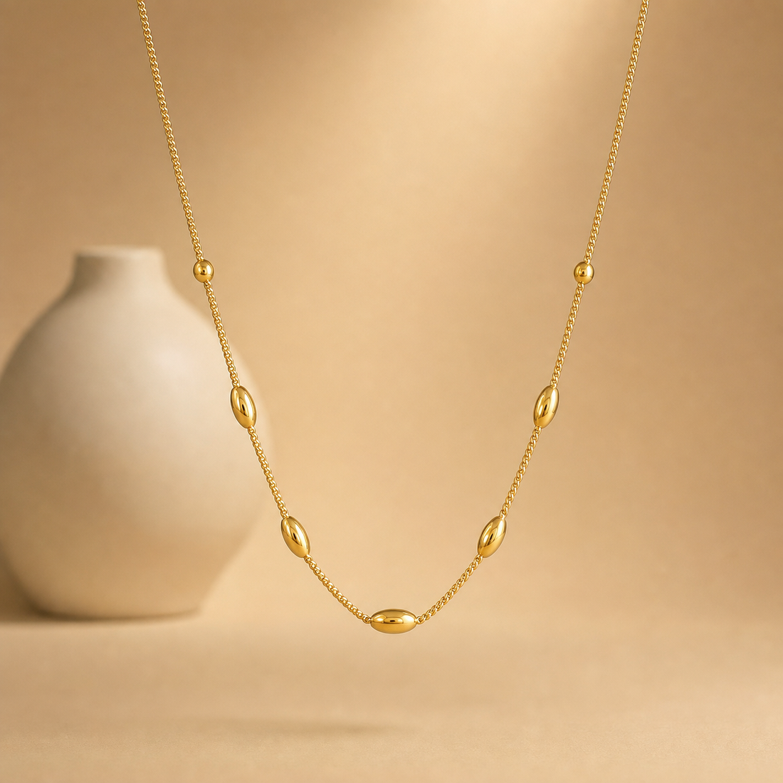 One necklace in yellow-gold-toned metal with a polished finish. A single strand of fine curb chain carries seven fixed solid-metal stations threaded directly onto the chain: five elongated oval rice-shaped beads, each with a small recessed detail on its face, and two smaller round beads, one on each side nearer the neck.