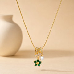 A single gold-tone pendant necklace with a polished finish. Construction is one continuous strand of fine snake chain, roughly 30 times the width of the largest component, with no visible extender; the closure is not visible in this view. The strand runs into a barrel-style pendant bail of stacked gold-tone rings channel-set with rows of small clear faceted stones, from which three jump-ring-mounted dangles hang: one side dangle is a small prong-set clear faceted round stone; the centre dangle is a flower