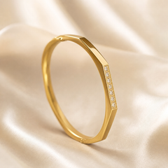 A single rigid bangle (kada) in yellow gold with a polished finish. The band is a continuous closed ring of medium gauge with an angular, faceted outer profile: broad flat bevels run along the band, giving a slightly hexagonal cross-section rather than a round one.