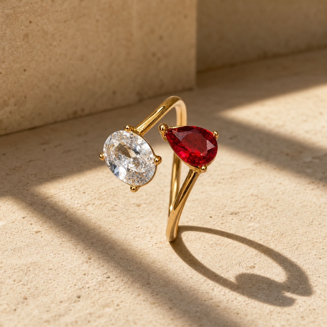 One finger-worn ring in polished yellow gold with a rounded, moderately slender band, roughly one-quarter the width of the head, formed as an open adjustable bypass band whose two ends each carry a stone. The head comprises two prong-set faceted stones facing one another: one oval-cut clear colourless stone and one pear-cut red stone of similar size, each held by four rounded prongs. The clear stone shows visible internal inclusions.