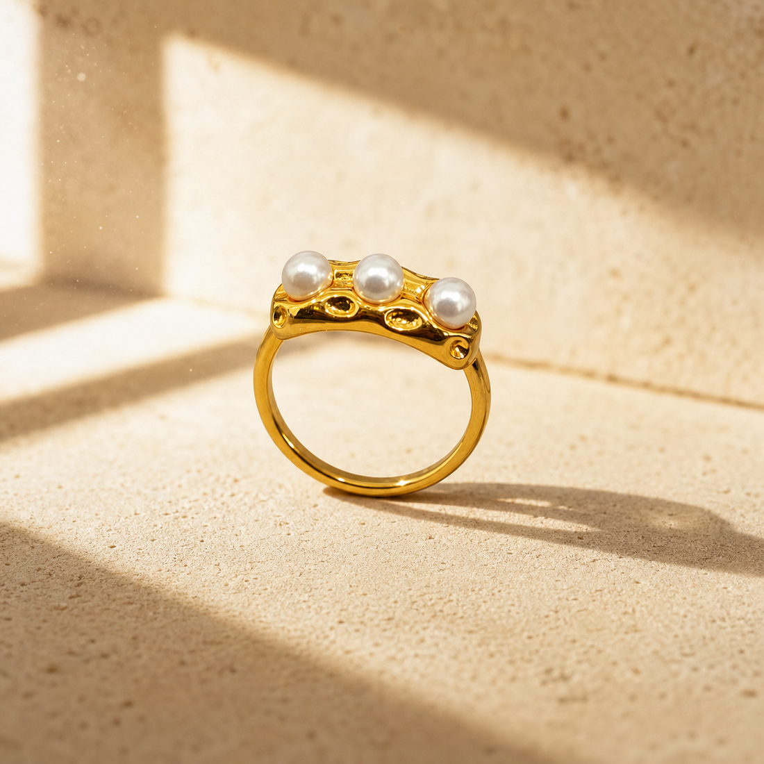One finger-worn ring in yellow-gold-toned metal with a polished, irregularly textured finish. The band is rounded in profile, a closed circle, roughly one-third the width of the head. The head is an elongated, organically shaped molten-style bar with a lumpy, hand-worked relief and several small recessed dimples across its face. Set into the head are three round white pearls of similar size, each seated in a low bezel-style cup, evenly spaced in a row.