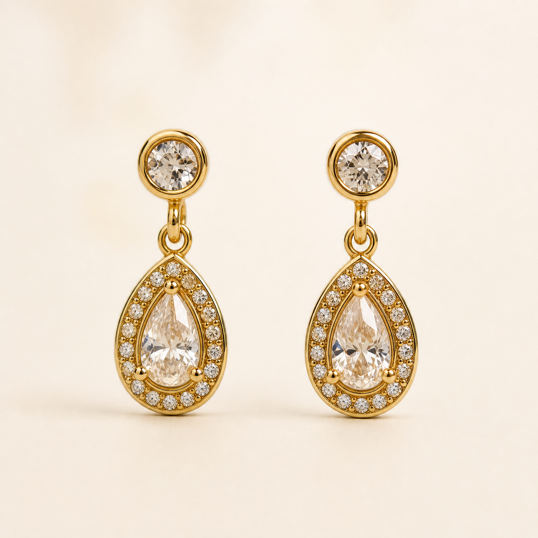 Two ear-worn earrings of one design in yellow-gold-toned metal with a polished finish. Each has a post fitting (back not visible, likely a standard push post), the post topped by a small round clear faceted stone in a smooth bezel. From the stud a short jump-ring link articulates to a teardrop-shaped drop section, giving two hinged sections; overall drop is roughly two and a half times the widest point (the teardrop).