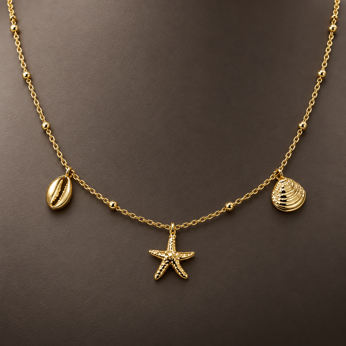 Gold-tone 316L stainless steel necklace with a bead-and-cable chain and three charms: a cowrie shell, a starfish and a ribbed shell, worn at the neck